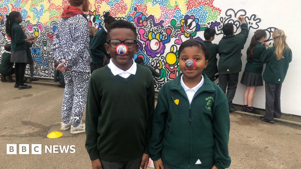 A man with red hair and beard is dressed in a black and white top, featuring cartoon drawings of a variety of characters. He is wearing one red nose with black and white doodle illustrations, and four of his fingers with a red nose with black and white doodle illustrations.