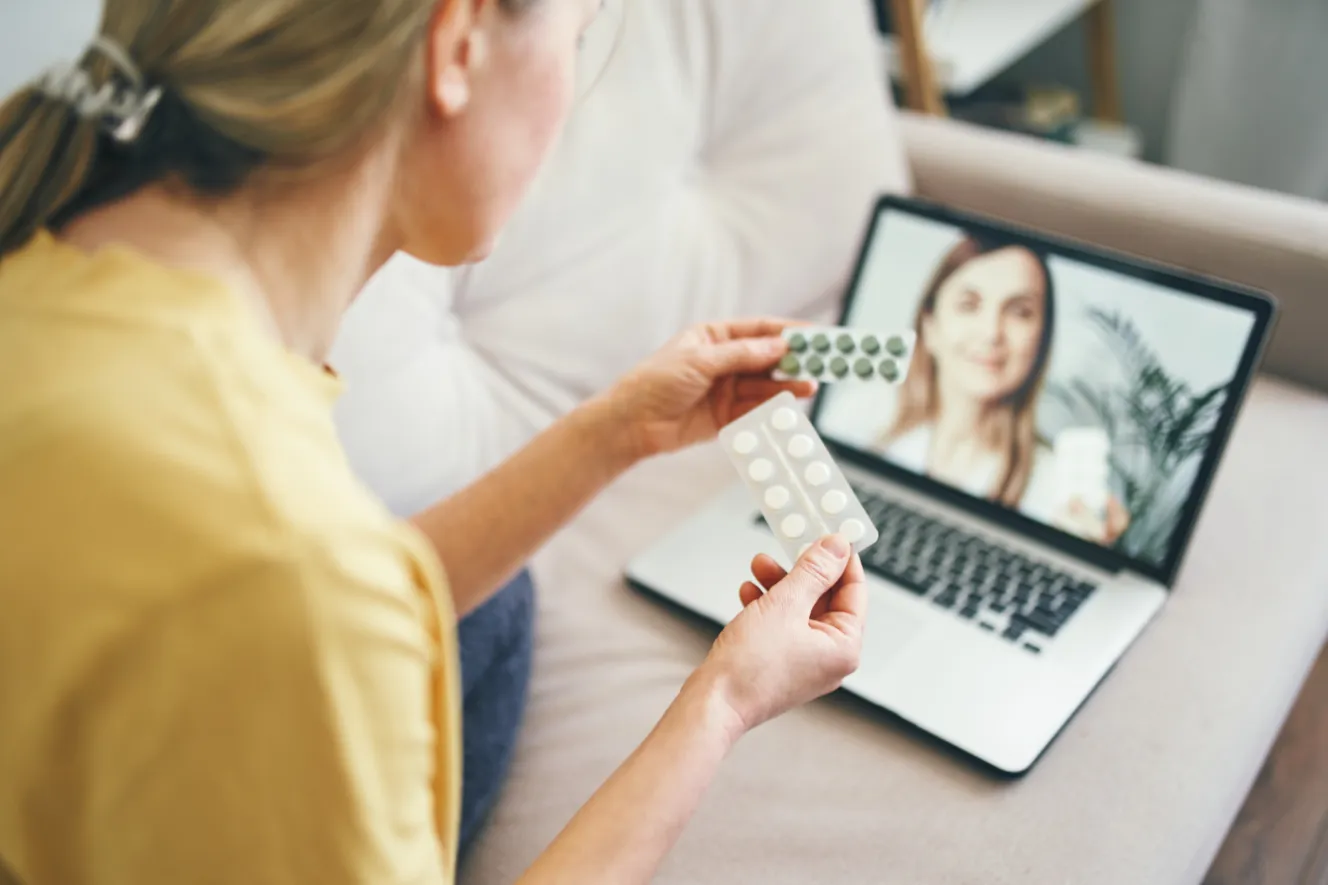 A woman holding two blister packs of pills, one green and one white, consults with a doctor on a laptop via video call.