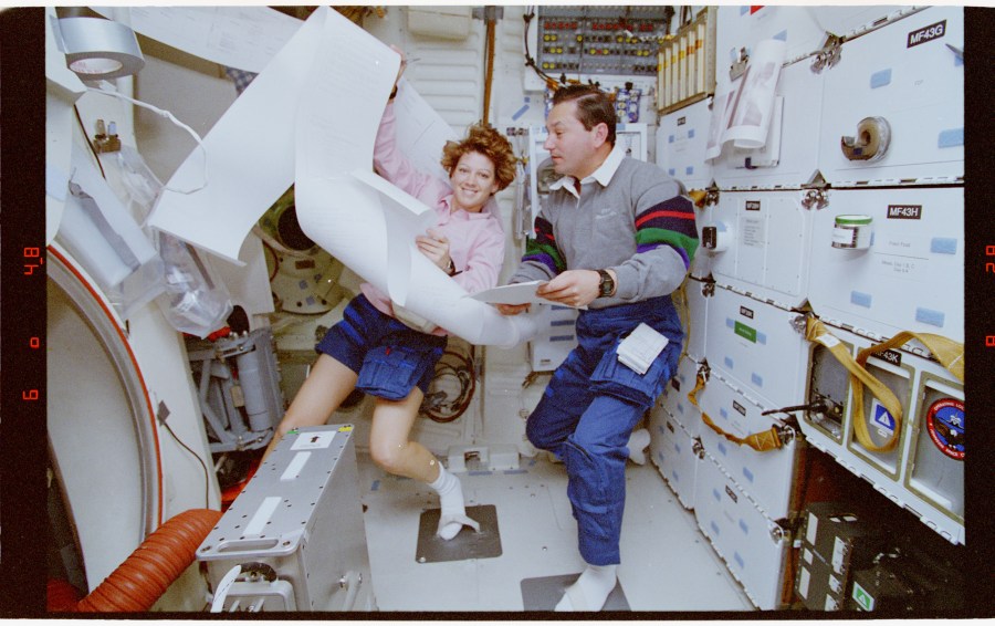 Pilot Eileen Collins and Mission Specialist Vladimir Titov posing with papers from the Thermal Impulse Printer System (TIPS) on the Space Shuttle Discovery's mid-deck.