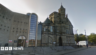 A general view of Kirkcaldy Sheriff Court. An older building made of dark stone is connected to a newer building, made of lighter stone, by several glass panels. A large brick archway is in the middle, which is also filled with glass panels.