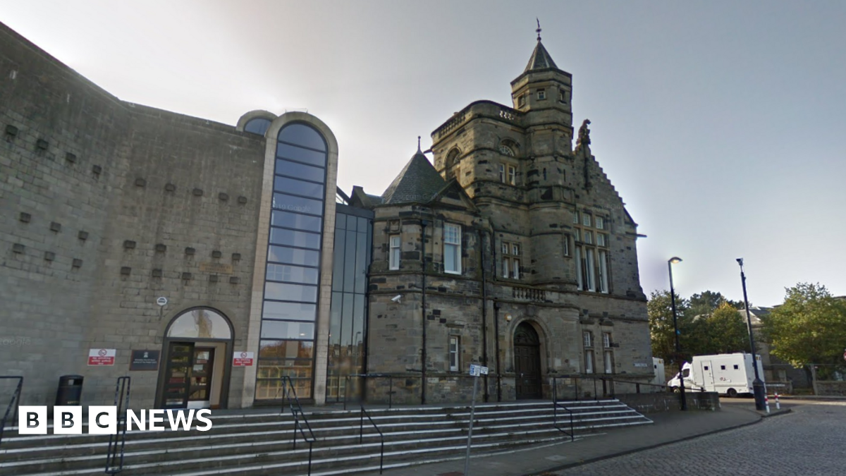 A general view of Kirkcaldy Sheriff Court. An older building made of dark stone is connected to a newer building, made of lighter stone, by several glass panels. A large brick archway is in the middle, which is also filled with glass panels.