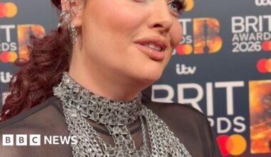 A woman on the red carpet, she has red hair tied back and a mesh top with huge chain necklaces.