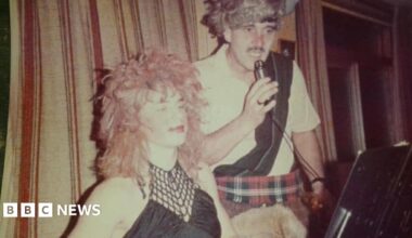 Barlow and Trevor Davies in fancy dress on stage. Barlow wears a long red wig and black woman's top, while Davies wears red tartan kilt, white shirt with a black sash, and a grey wig. Davies holds a microphone, with a stage curtain behind them both.