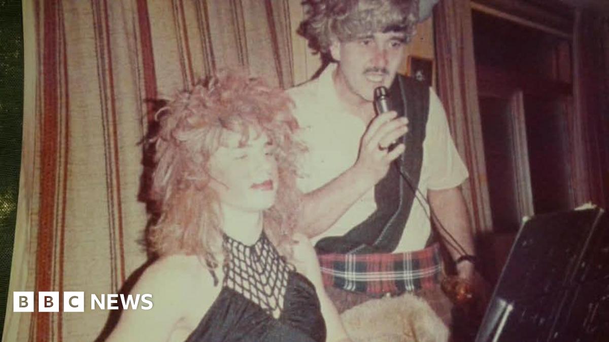Barlow and Trevor Davies in fancy dress on stage. Barlow wears a long red wig and black woman's top, while Davies wears red tartan kilt, white shirt with a black sash, and a grey wig. Davies holds a microphone, with a stage curtain behind them both.