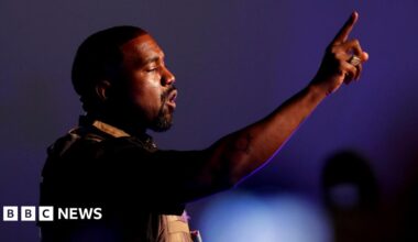 Kanye West addresses a crowd. He is a black man with a dark goatee and is wearing a military style vest. He is holding one finger up as he speaks on a stage.