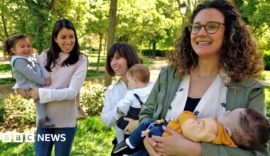 Tania has brown hair and is holding her baby in a sunny park.