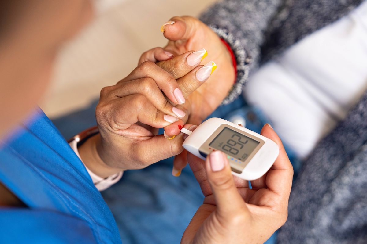 Home healthcare for seniors with diabetes. Female nurse in blue uniform measures blood sugar using a glucometer on an elderly woman resting in bed. Concept of health, chronic care, and senior well-being.