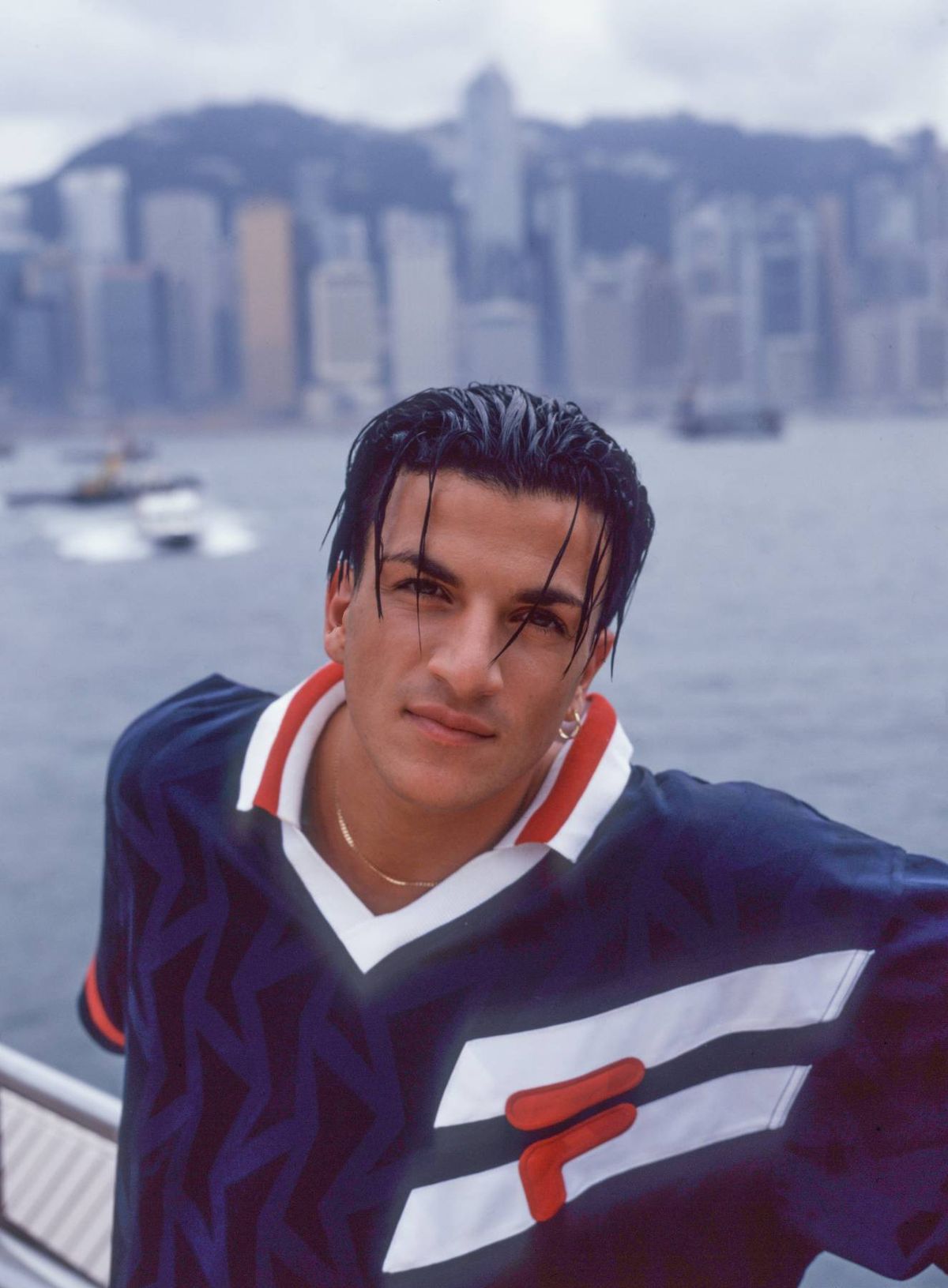 An individual with a serious expression, dressed in a blue and white athletic shirt, stands by a body of water. The background features a cityscape with boats and buildings.