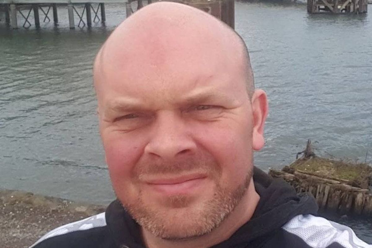 An individual, dressed in a black and white hoodie, is standing near a waterfront, smiling at the camera with a backdrop of a pier and waterbody.