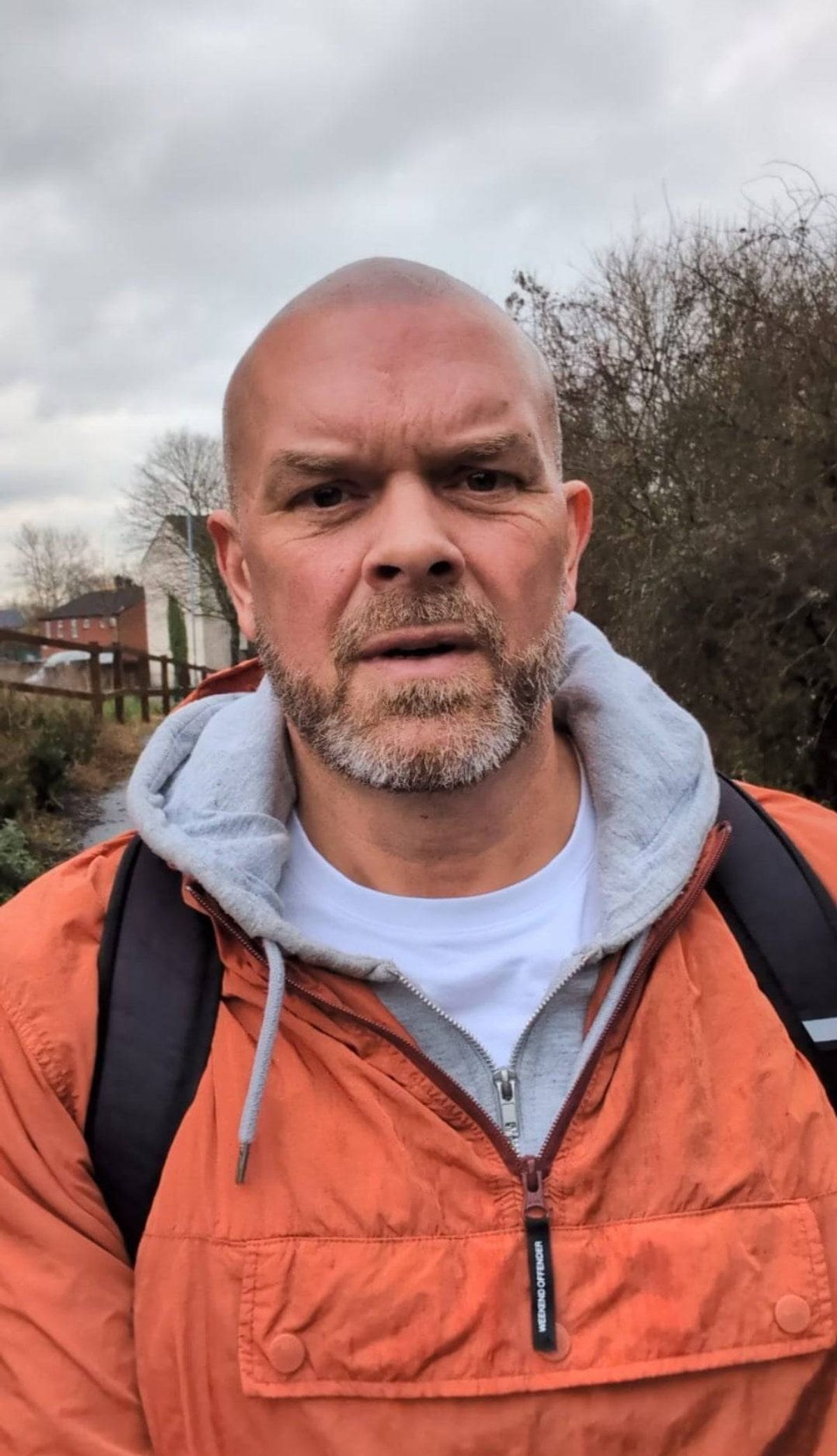 An individual stands outside during the daytime, clad in an orange jacket with a gray hood and a black backpack. The individual's facial expression appears serious, and the backdrop includes greenery and a cloudy sky.