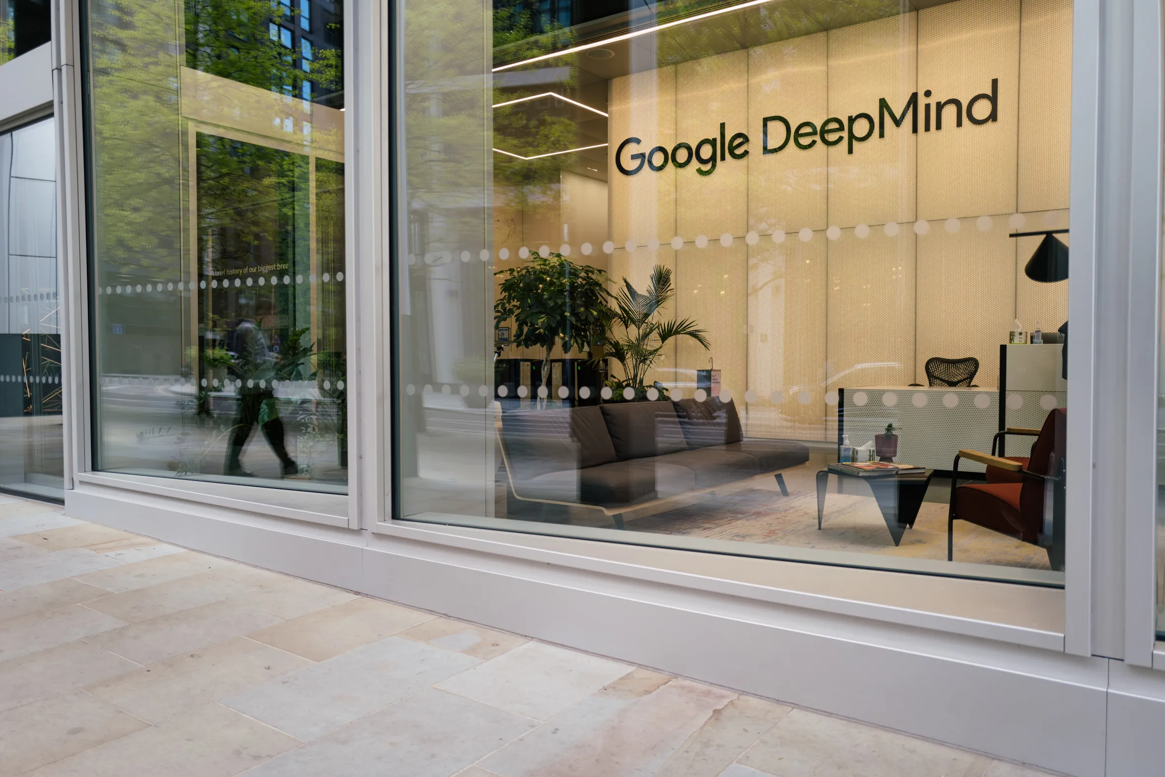 A Google DeepMind sign visible through the window of a building, showing the reception area with a sofa, desk, and plants.