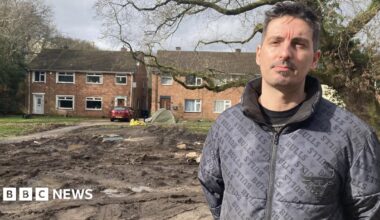 local resident Pedro Oliveira is wearing a black jacket and is standing in front of some muddy waterlogged land, in front of some beige-bricked houses.