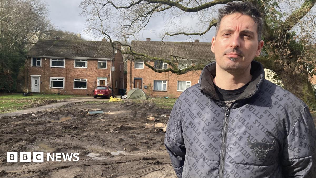 local resident Pedro Oliveira is wearing a black jacket and is standing in front of some muddy waterlogged land, in front of some beige-bricked houses.