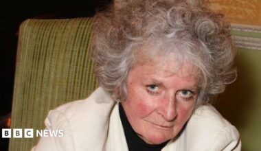 Maggi Hambling looks at the camera and slightly smiles as she leans to the right. She sits on a green sofa. She has short grey curly hair and wears a white jacket with a black top underneath.