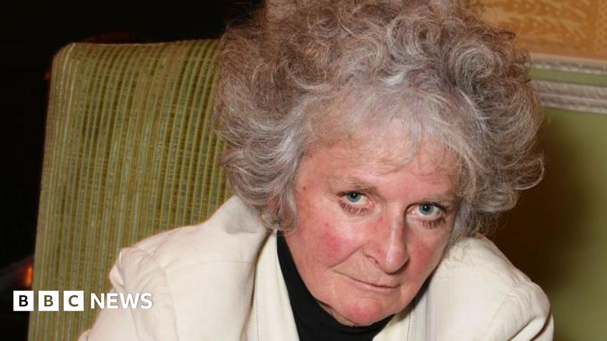 Maggi Hambling looks at the camera and slightly smiles as she leans to the right. She sits on a green sofa. She has short grey curly hair and wears a white jacket with a black top underneath.