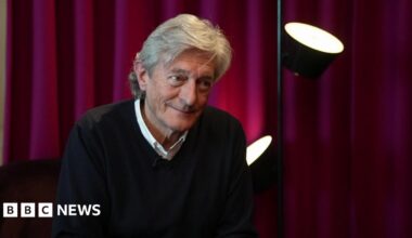 An older man with a checked scarf and grey hair sits in a radio studio with headphones on