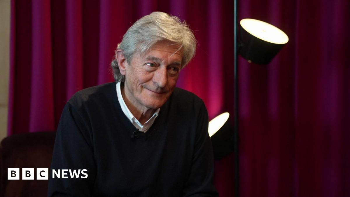 An older man with a checked scarf and grey hair sits in a radio studio with headphones on