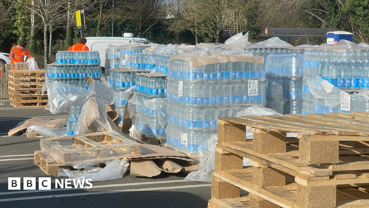 Stacks of bottled water set up in a car park, next to empty pallets.