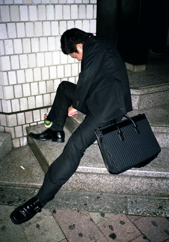 A man in a black suit is slouched and sitting awkwardly on outdoor steps at night, with his head down and a large black bag resting on his leg. The scene suggests exhaustion.
