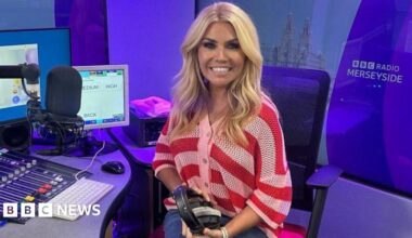 Leanne Campbell, who has wavy blonde hair and is wearing a chunky knit red and white striped cardigan, sits in a swivel chair by a studio desk with a large presenter's microphone to her right. She is holding a pair of black earphones.