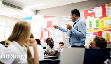 A stock image shows a secondary teacher standing teaching in a classroom wearing a suit and tie. The students are looking at him from their desks, wearing white short sleeved shirts.