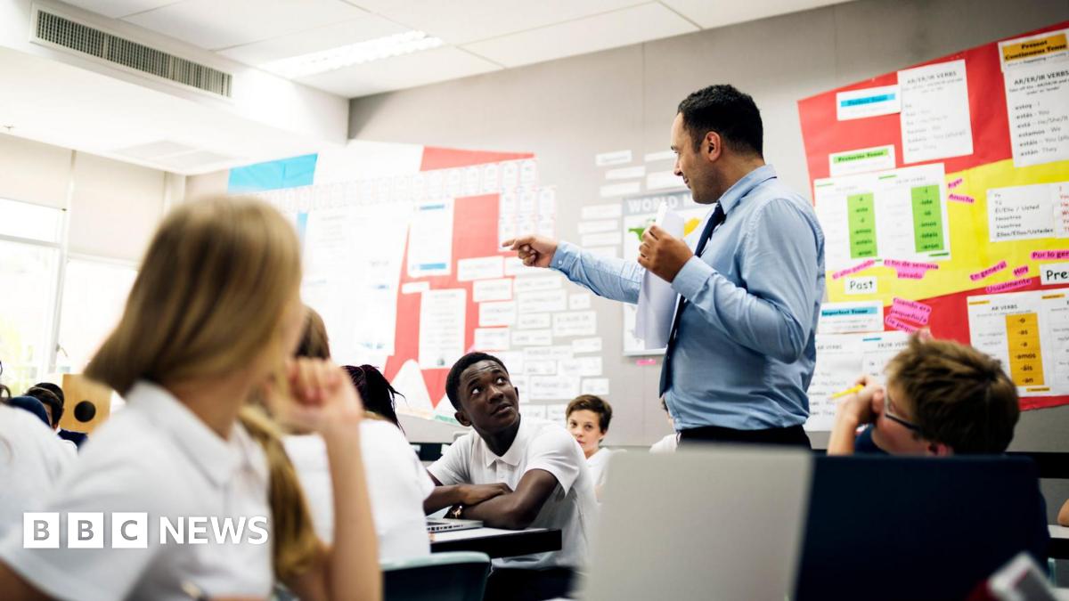 A stock image shows a secondary teacher standing teaching in a classroom wearing a suit and tie. The students are looking at him from their desks, wearing white short sleeved shirts.