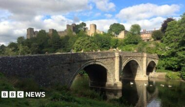 Ludlow Castle and Dinham bridge pictured on a sunny day. Swans can be seen in the river and people crossing the bridge