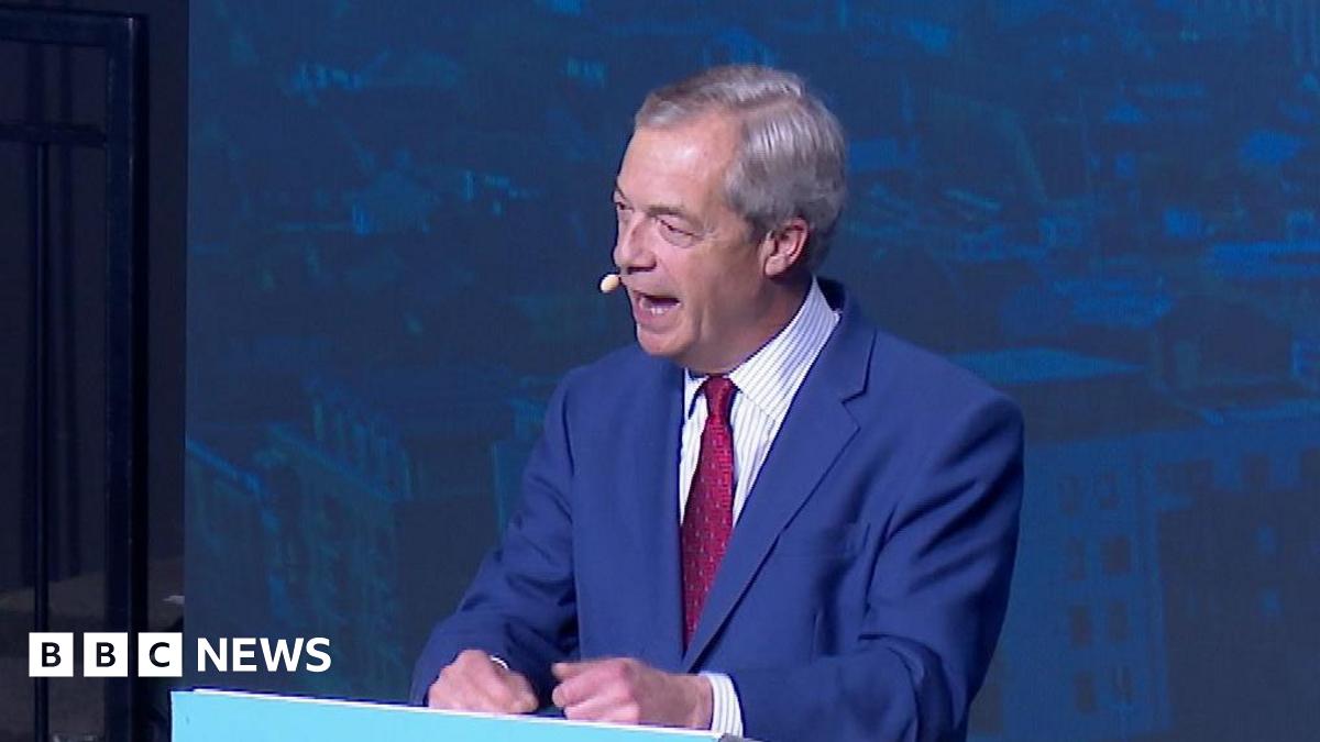 Nigel Farage stands at a powder blue podium and is speaking on stage in a blue suit and red tie. There is a blue backdrop behind him.