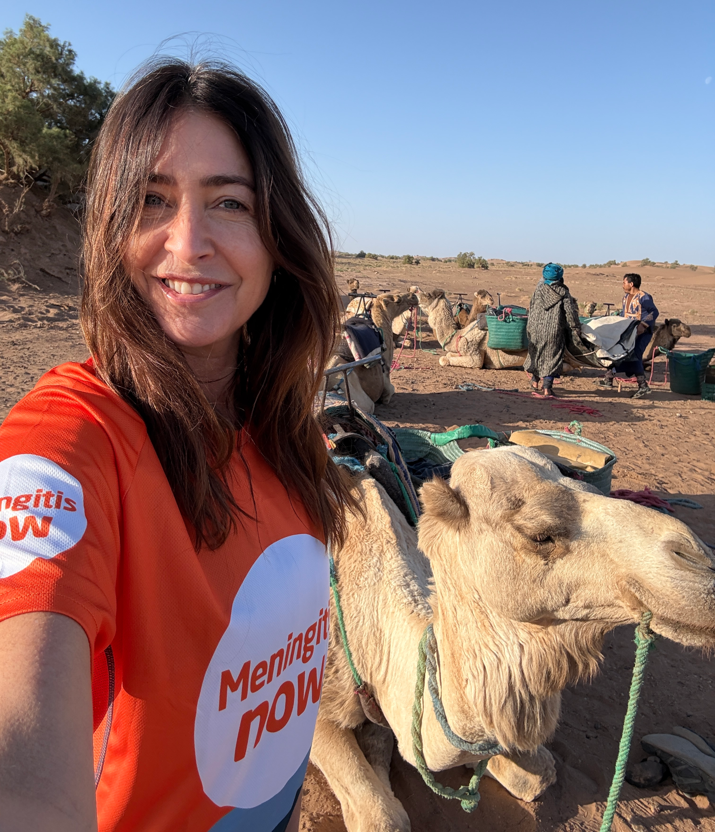 A woman in an orange "Meningitis Now" shirt smiles next to a camel, with more camels and people in the desert background.