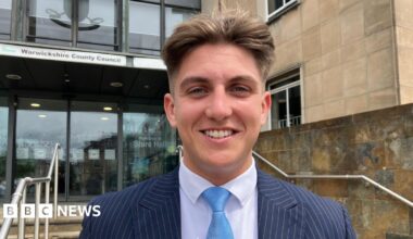 George Finch is looking directly at the camera smiling. He is wearing a navy pinstriped suit with a white shirt and a light blue tie. He is standing outside of the entrance to a building, in the background some steps and handrails can be seen, and behind them some automatic doors, above which is a sign that says Warwickshire County Council.