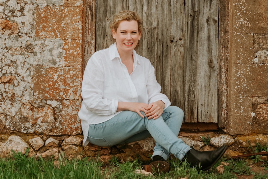 A smiling blonde woman sits on the stone step of a rustic-looking building.
