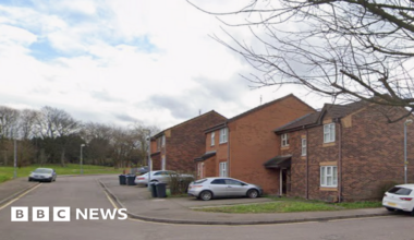 A woman in her 40s is sitting on a sofa in her living room holding a walking stick. She has long orange hair. Behind her is cushions and lighting.
