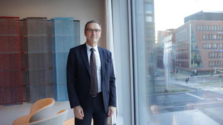 Silmälasipäinen mies seisoo avarassa aulassa suuren ikkunan edessä.