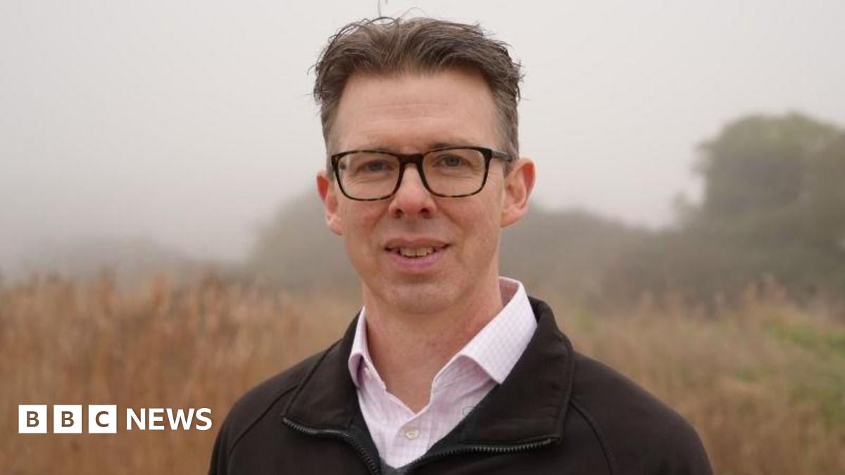 Eliot Lyne standing on the Cley Marshes nature reserve. He has short brown hair and is wearing glasses. He is wearing a green top and white shirt. There are plants and shrubs behind him, which are out of focus.