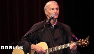 Len Garry is pictured on stage with grey hair and wearing a black shirt and playing an acoustic guitar