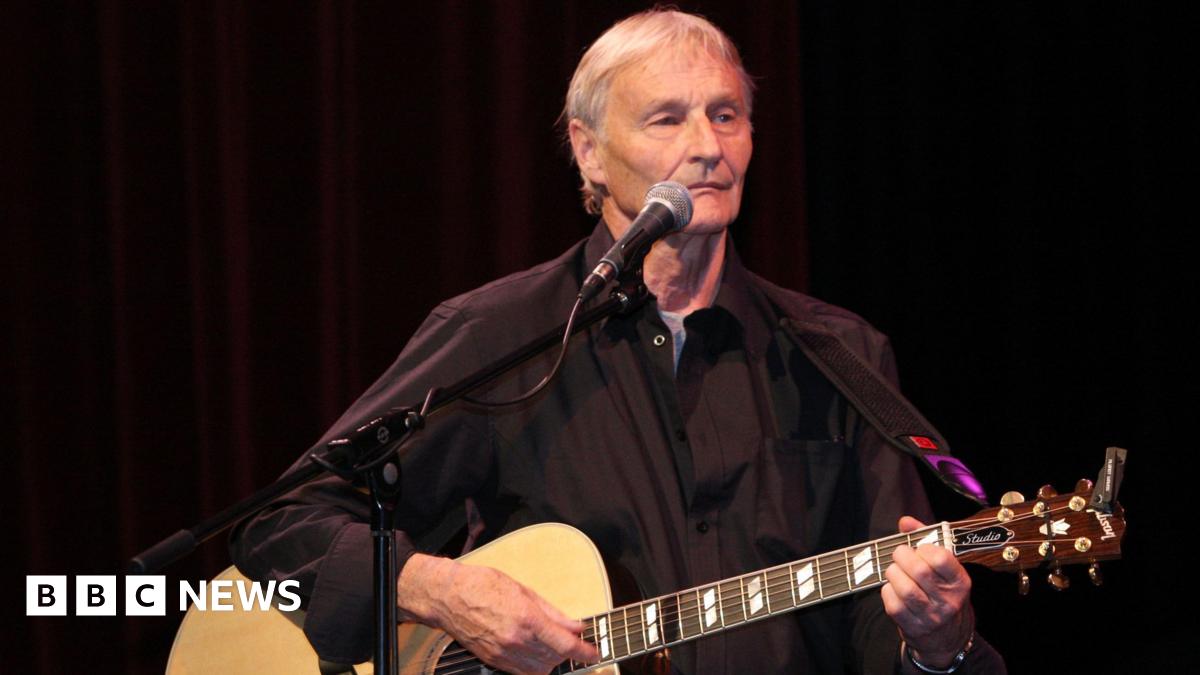 Len Garry is pictured on stage with grey hair and wearing a black shirt and playing an acoustic guitar