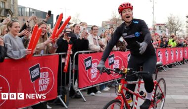 Radio 1 DJ Greg James riding a tandem bicycle into Keel Square, Sunderland, in front of a crowd of cheering people. He is wearing black cycling clothes. There are red banners for Comic Relief Red Nose Day covering silver crowd barriers.