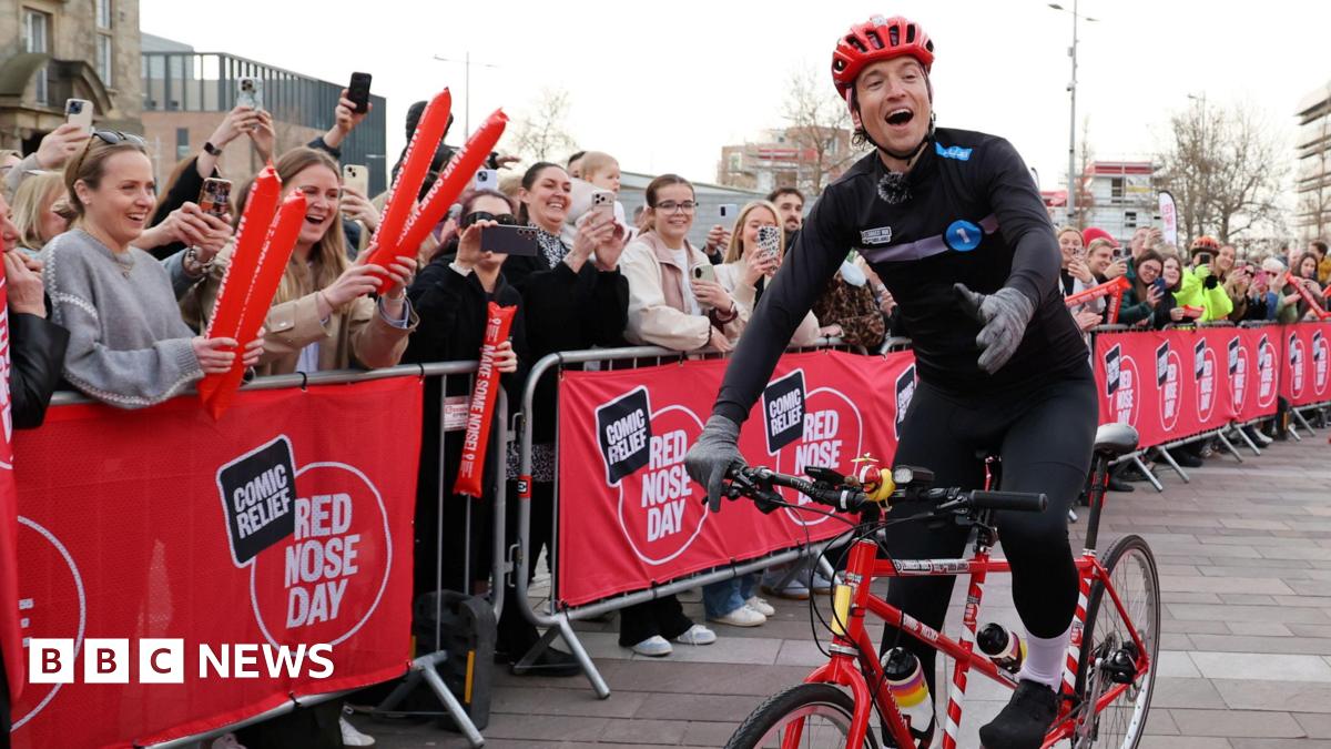 Radio 1 DJ Greg James riding a tandem bicycle into Keel Square, Sunderland, in front of a crowd of cheering people. He is wearing black cycling clothes. There are red banners for Comic Relief Red Nose Day covering silver crowd barriers.