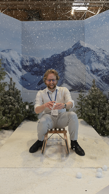 Man sitting on a small wooden stool in a staged snowy mountain scene, smiling as artificial snow falls around him, with fir trees and a large alpine backdrop behind.