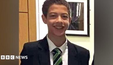Noah Donohoe, wearing a white shirt, black and green tie, and a black suit jacket. He has short brown hair and is smiling at the camera.