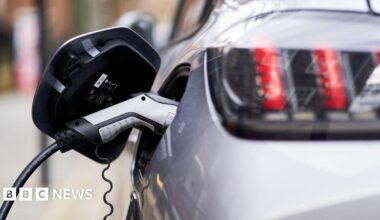 A close up image of a silver and black coloured electric vehicle charger which is plugged into a silver car. The car is out of focus but you can see the rear of the vehicle and the back headlights.