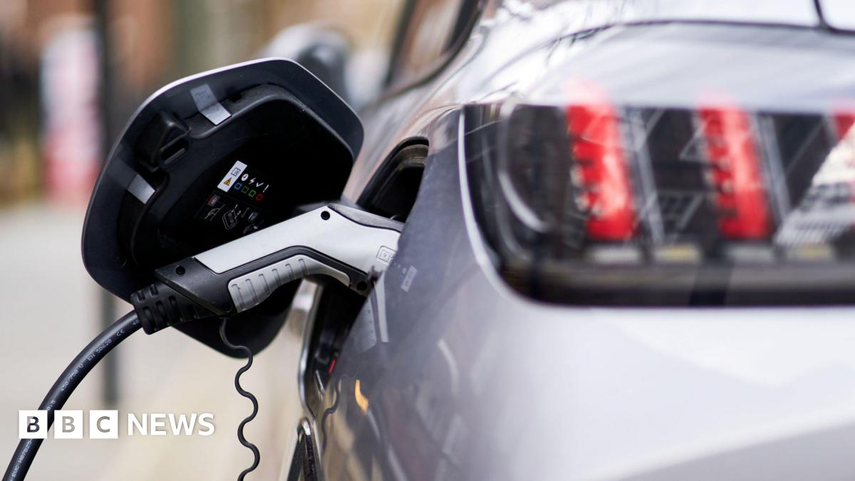 A close up image of a silver and black coloured electric vehicle charger which is plugged into a silver car. The car is out of focus but you can see the rear of the vehicle and the back headlights.
