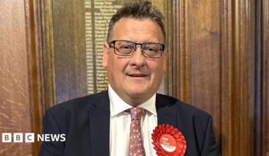 Karl Turner the succesful Labour candidate for Hull East pictured on election night in 2024 wearing a dark suit jacket, white shirt, and patterned pink tie, standing indoors against a wooden paneled wall. A large red rosette with the word ‘Labour’ and smaller text is pinned to the left side of the jacket.