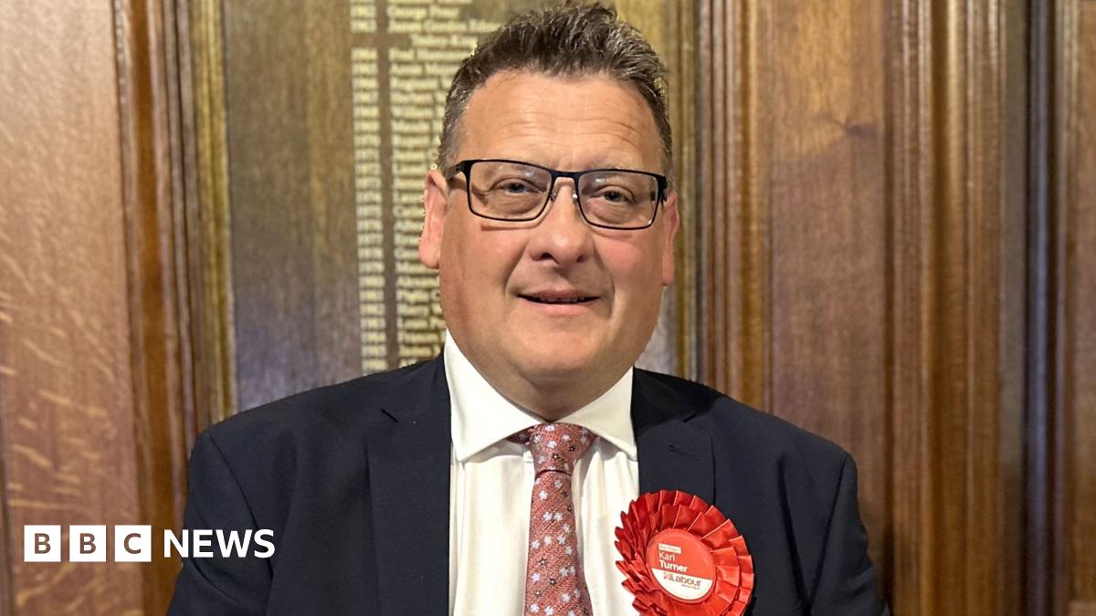 Karl Turner the succesful Labour candidate for Hull East pictured on election night in 2024 wearing a dark suit jacket, white shirt, and patterned pink tie, standing indoors against a wooden paneled wall. A large red rosette with the word ‘Labour’ and smaller text is pinned to the left side of the jacket.