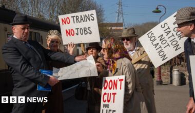 A group of protestors dressed in 1960s clothing carrying placards, harrassing a man dressed as Dr Beeching, who is looking at a railway map of the UK.