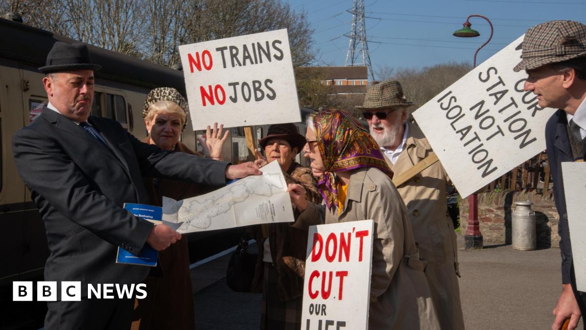 A group of protestors dressed in 1960s clothing carrying placards, harrassing a man dressed as Dr Beeching, who is looking at a railway map of the UK.