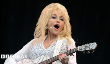 Dolly Parton wearing a sheer white blouse with balloon sleeves and white trousers holding a white and silver acoustic guitar on stage singing into a microphone attached to her head.