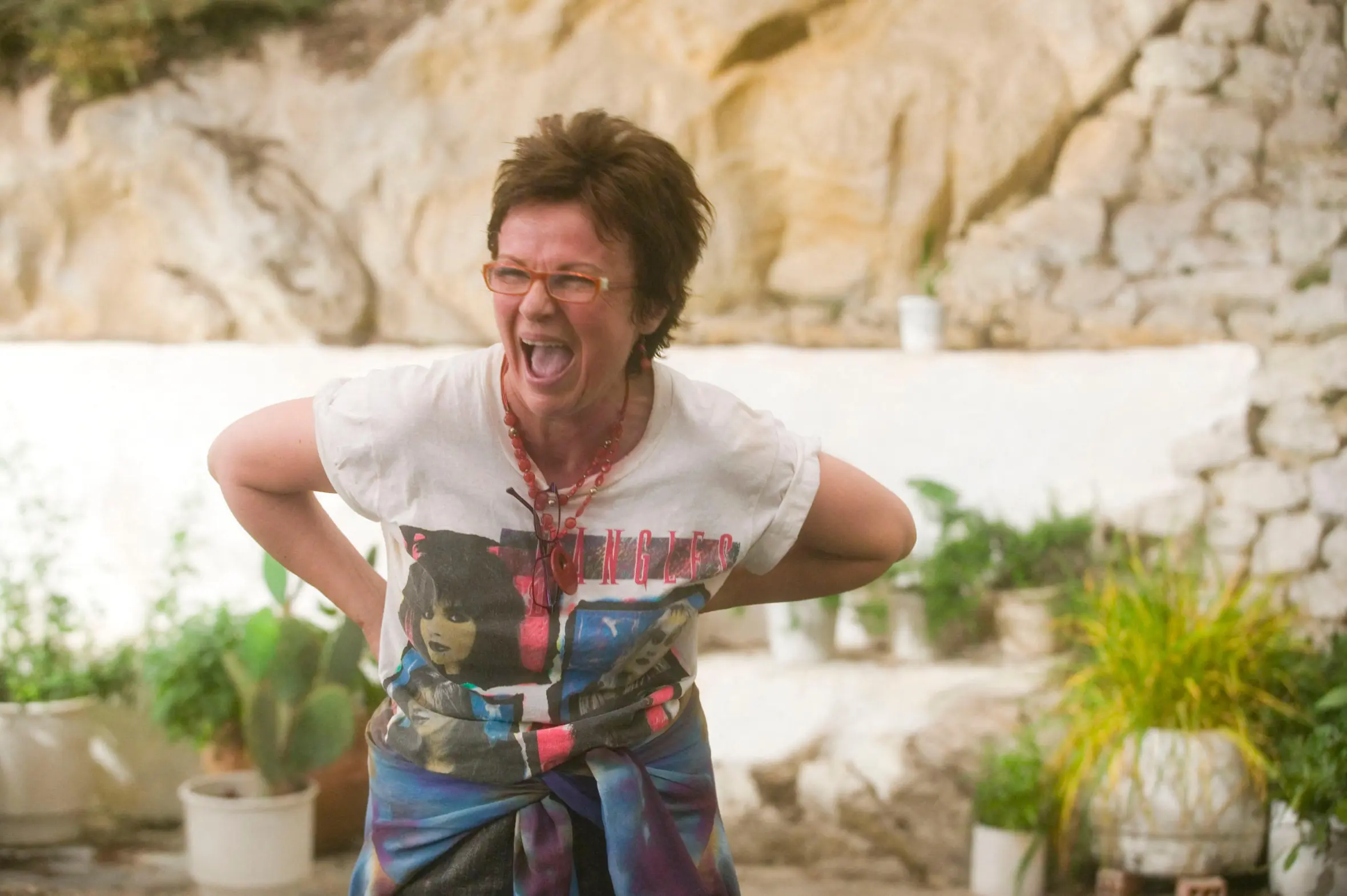 Julie Walters laughing with her hands on her hips, wearing a white t-shirt and orange glasses, in a scene from the movie "Mamma Mia!"