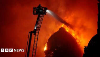 A firefighter on a large ladder sprays water over a burning domed building
