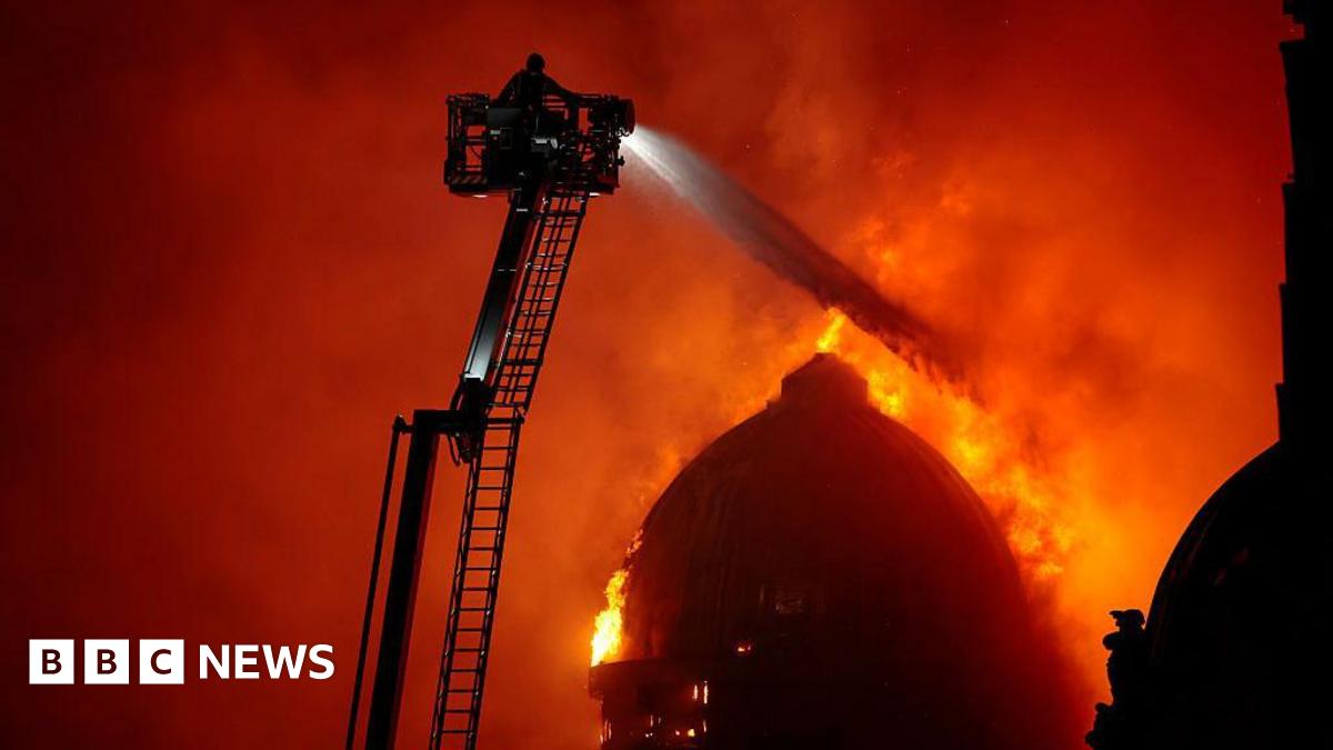 A firefighter on a large ladder sprays water over a burning domed building